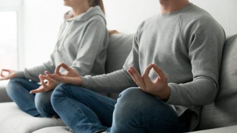 Calm couple meditating together at home on sofa, mindful peaceful man and woman practicing yoga sitting in lotus pose on couch in living room, focus on hands in mudra circle, close up view