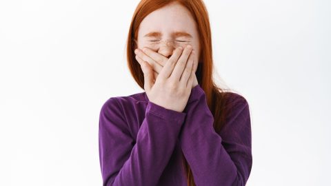 Portrait of cute redhead girl sneezing, covering her mouth and wrinkle nose, standing over white background. Kid hiding her smile, laughing and joking.