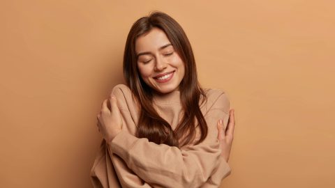 I love myself. Pretty smiling woman embraces herself gently, expresses self love, keeps eyes closed with pleasure, feels comfortable and fullfilled, being egoistic person, isolated on brown background