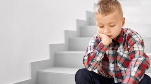 One sadly school boy in checked shirt sitting on stairs at corridor of school and crying. Little school boy leaning head by hands and thinking. Concept of bed marks at school.